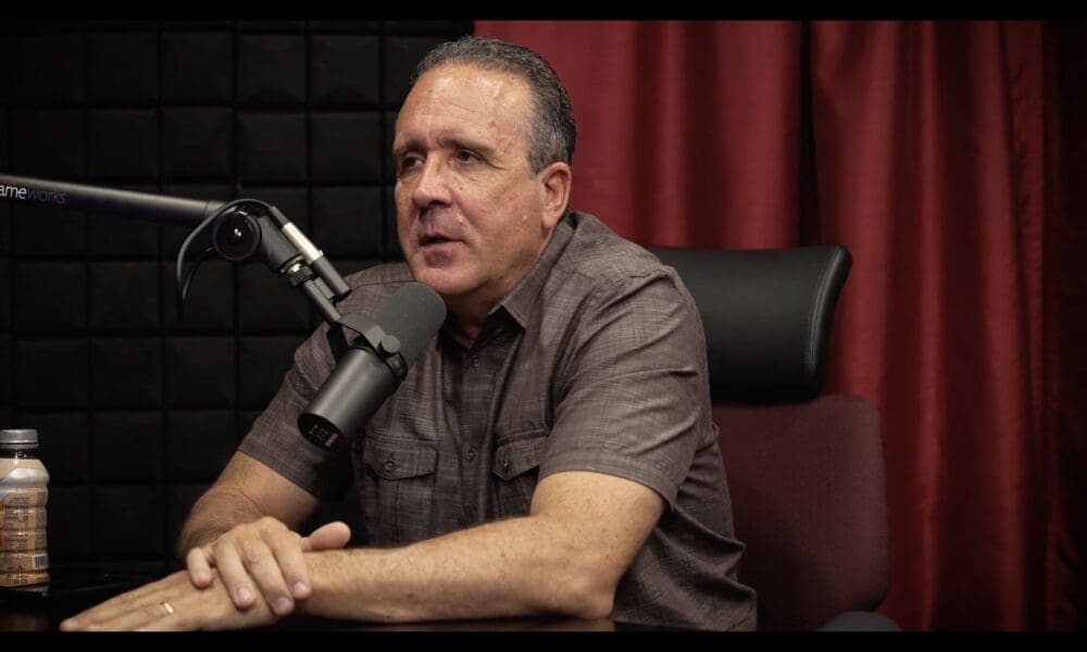 man in gray shirt in podcast studio with red curtain behind him